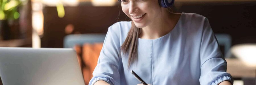 smiling woman wearing headphones taking notes while attending an online meeting - learning management system​